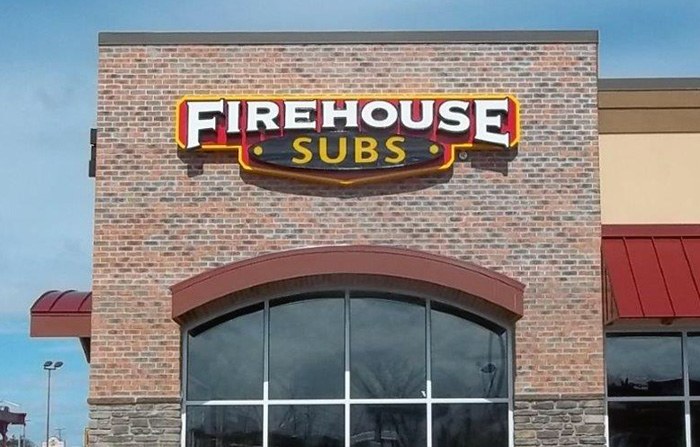 A Firehouse Subs storefront sign mounted on a brick building exterior, with the logo in red, yellow, and black lettering, red awnings below, and a clear blue sky above.
