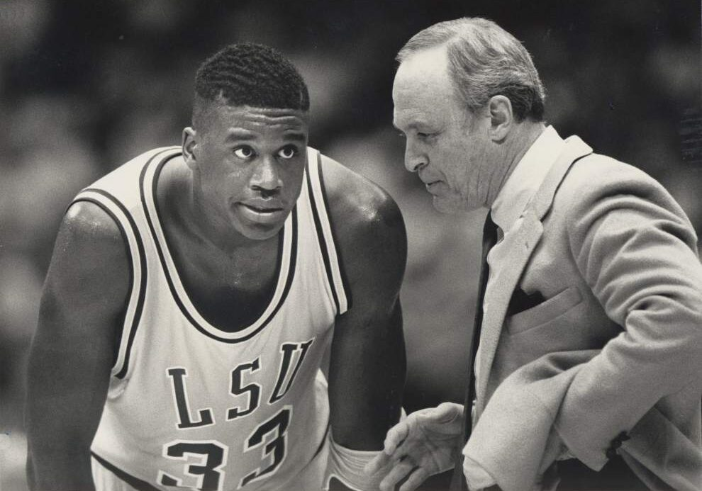 Black-and-white archival photo of a young Shaquille O'Neal in an LSU #33 jersey talking with coach Dale Brown courtside