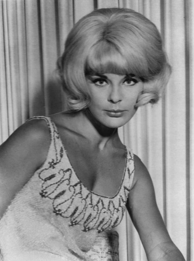 A black-and-white glamour portrait of a young woman with voluminous bouffant hair and a beaded gown, looking at the camera.