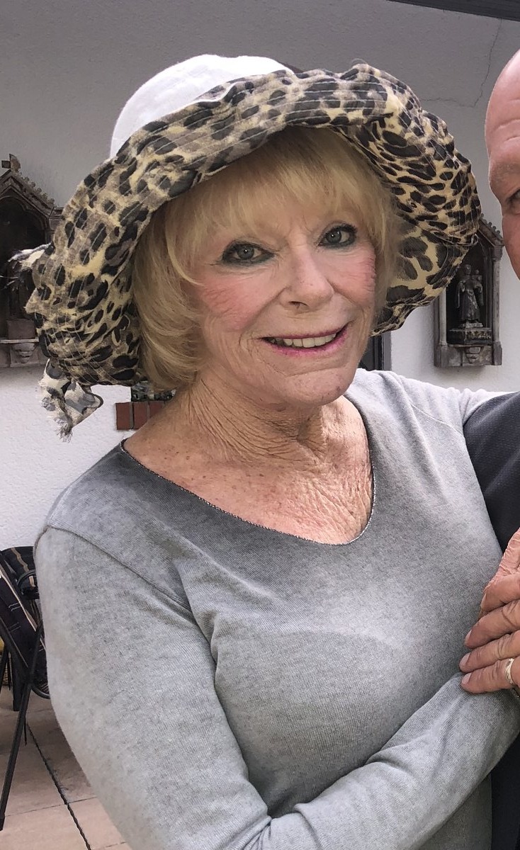 An older woman with blonde hair wearing a leopard-print hat, smiling broadly.