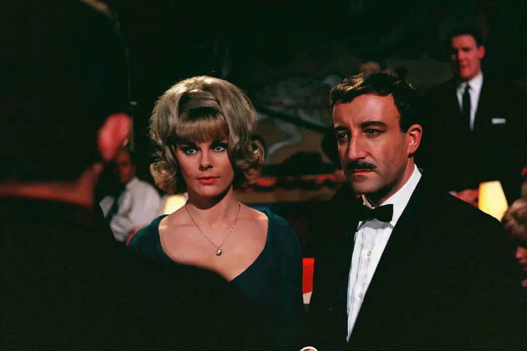 A color film still from 'A Shot in the Dark' (1964) showing Elke Sommer, as Maria Gambrelli, and Peter Sellers, as Inspector Clouseau in a tuxedo with a mustache, standing together at a formal event with other guests visible in the background.